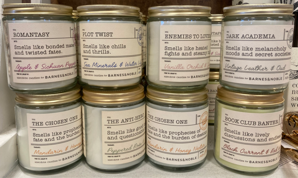 A photograph of candles in jars for sale. There are two rows of four candles each (one row stacked on the other row). The candles appear to be white in color. The jars are mostly covered with labels designed to resemble bookplates with names like "Romantasy: Smells like bonded mates and twisted fates" and "Dark Academia: Smells like melancholy moods and secret societies". Each label also includes an aroma description.